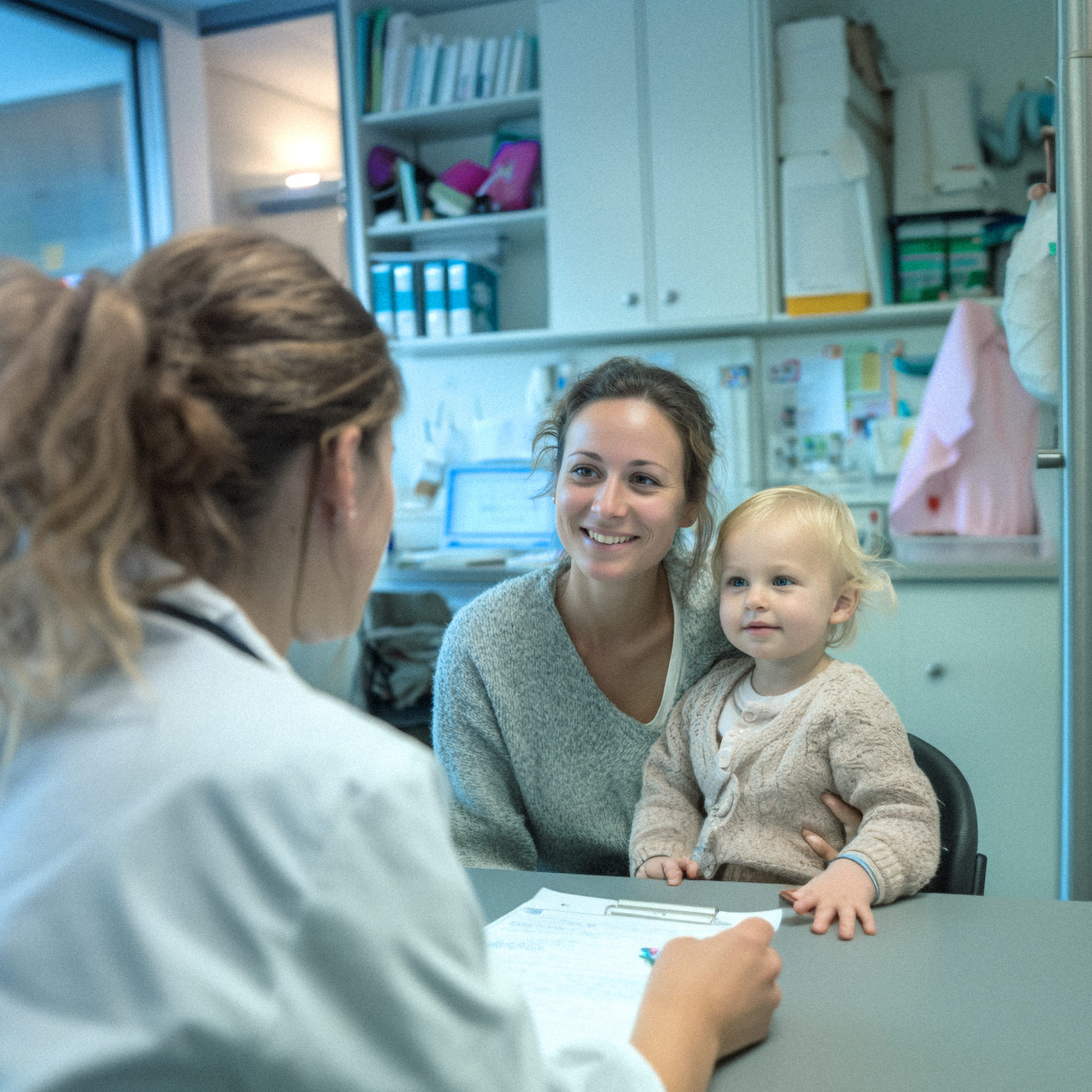 Eine lächelnde Mutter sitzt mit ihrem Kleinkind in einem Behandlungszimmer der Ostseeklinik Grömitz einer Ärztin gegenüber, die im Vordergrund angeschnitten zu sehen ist.