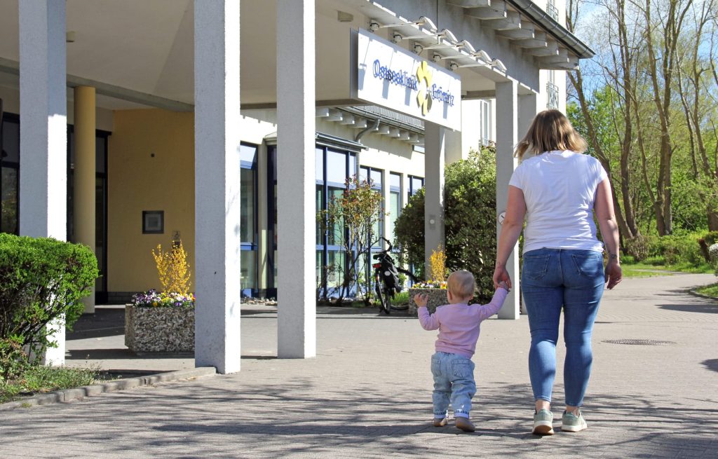 Eine Mutter und ein kleines Kind gehen Hand in Hand auf den Eingangsbereich der Ostseeklinik Grömitz zu. Im Hintergrund ist das Klinikgebäude mit dem blau-gelben Logo über dem Eingang und einer gepflegten Außenanlage unter strahlend blauem Himmel zu sehen.