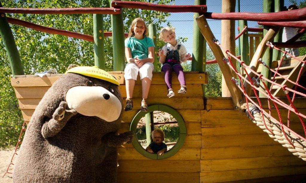 Zwei Kinder sitzen auf einem hölzernen Spielschiff im Außenbereich der Ostseeklinik Grömitz, während das Klinikmaskottchen Grömel, ein großer brauner Bär mit gelber Kappe, daneben steht und winkt.