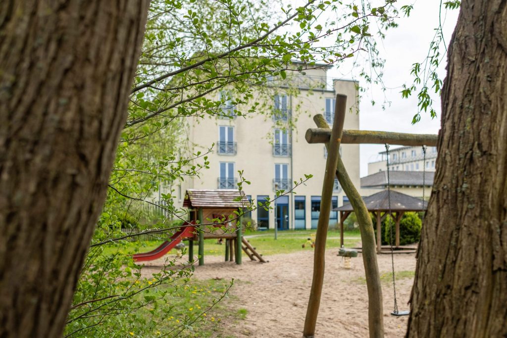 Kinderspielplatz im Außenbereich der Ostseeklinik Grömitz mit einer Rutsche, Schaukeln und einem Pavillon auf einer Sandfläche, im Hintergrund ist das Klinikgebäude mit hellgelber Fassade und französischen Balkonen zu sehen.