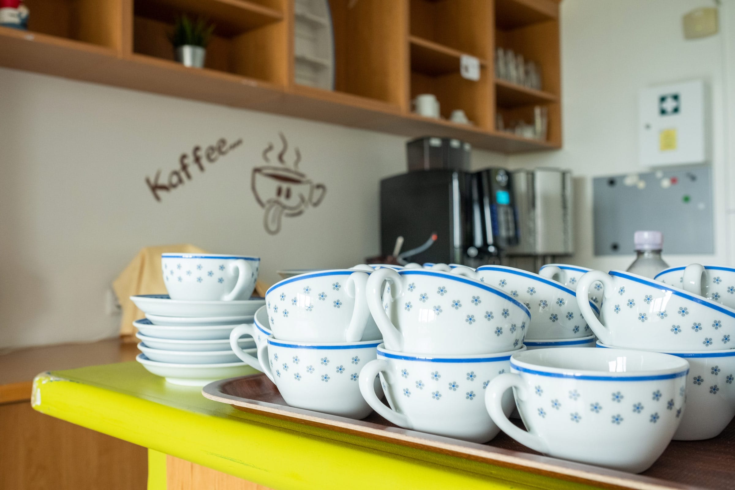 Weiße Porzellantassen mit blauen Gänseblümchen-Mustern und blauem Rand, gestapelt auf einem Tablett. Im Hintergrund eine Wand mit der Aufschrift "Kaffee" und einer Kaffeetassen-Grafik sowie eine moderne Kaffeemaschine.