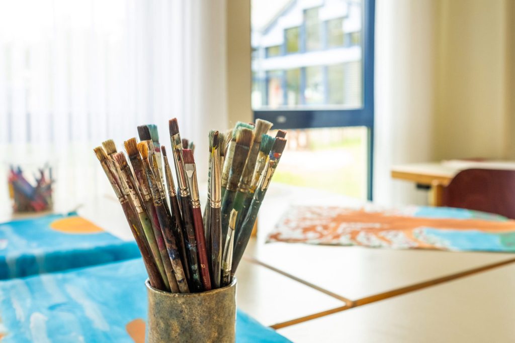 Im hellen Kreativraum: Nahaufnahme eines Bechers mit vielen genutzten Pinseln in, im Hintergrund sind Tische mit Malereien und ein großes Fenster erkennbar.