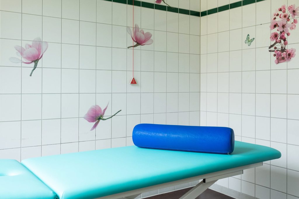 Heller Behandlungsraum in der Ostseeklinik Grömitz mit einer türkisfarbenen Untersuchungsliege, einer blauen Lagerungsrolle und floralen Wanddekorationen.