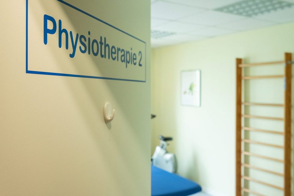 Schild „Physiotherapie 2“ an einer offenen Türe in der Ostseeklinik Grömitz mit Blick in einen Behandlungsraum, in dem ein Ergometer und eine Sprossenwand zu sehen sind.