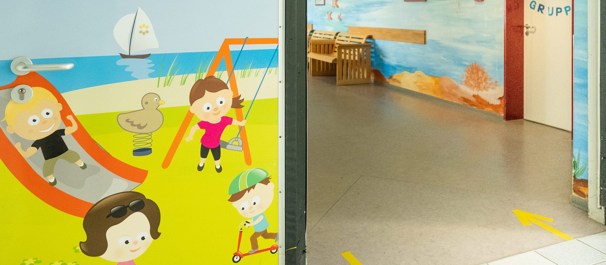 Blick in einen freundlichen Flur der Ostseeklinik Grömitz mit einer bunten Illustration spielender Kinder links an der Tür und dem Flur rechts mit einem Wandgemälde eines Strandes.