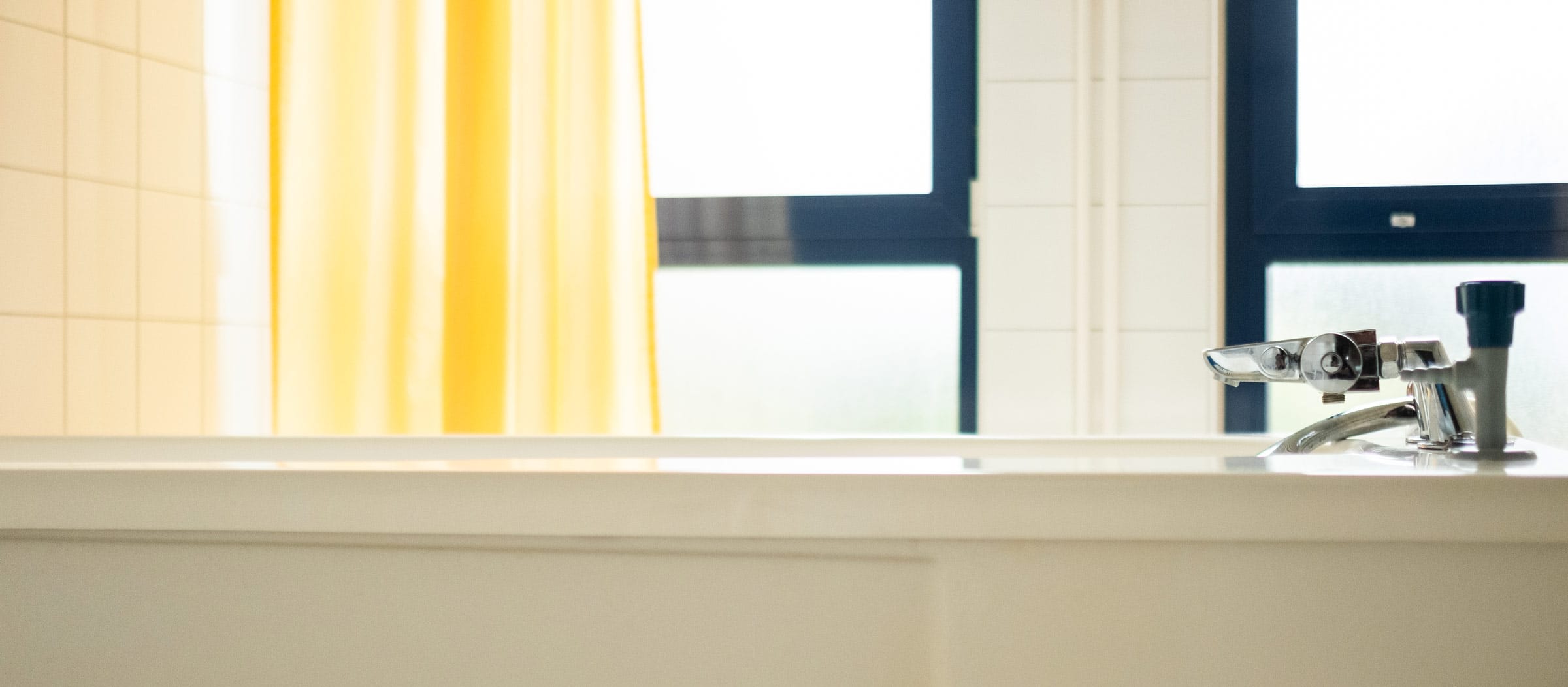 Hell gestalteter Therapieraum in der Ostseeklinik Grömitz mit einer weißen Therapiewanne mit Chrom-Armatur im Vordergrund und einem sonnengelben Vorhang und blickdichten Fenstern Hintergrund.
