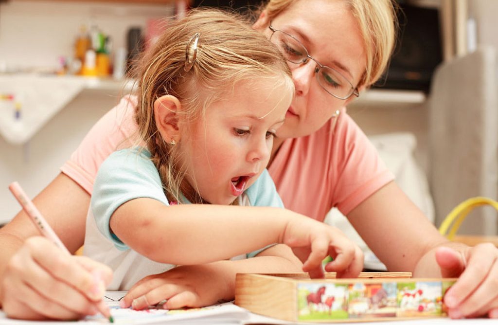 Ein junges Mädchen mit blonden Haaren sitzt neugierig an einem Tisch und greift in eine kleine Holzkiste, während seine Mutter ihr über die Schulter schaut und sie aufmerksam unterstützt.